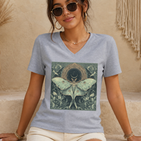 A woman wearing a light heather gray V-neck t-shirt with a large square graphic of a green luna moth and a crescent moon. She is wearing round sunglasses, gold hoop earrings, and a delicate gold necklace, sitting against a textured beige wall with boho decor.