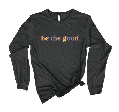 Flat lay long sleeve dark grey tee with graphic which reads “Be The Good” in colorful letters.