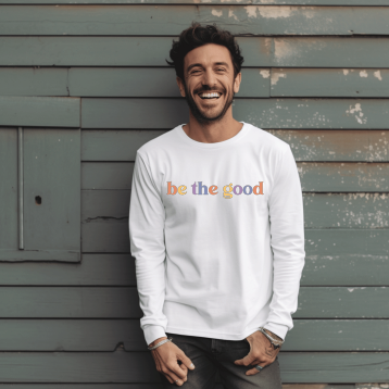 Young man wearing long sleeve white tee with graphic tha reads “Be The Good” in colorful letters.