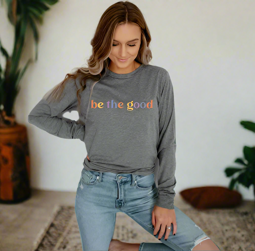 Young woman  long sleeve grey tee with graphic which reads “Be The Good” in colorful letters.