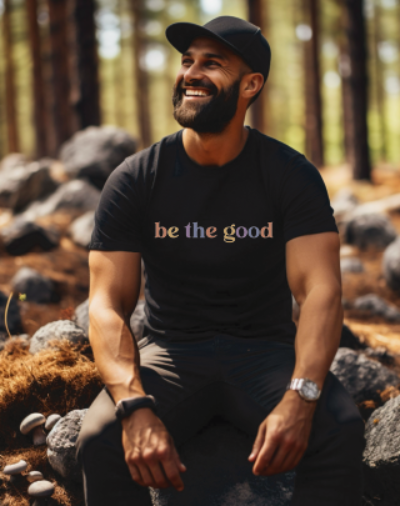 Young man in short sleeve black tee with graphic which reads “Be The Good” in colorful letters.