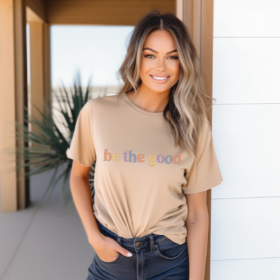Young woman in short sleeve tan tee with graphic which reads “Be The Good” in colorful letters.