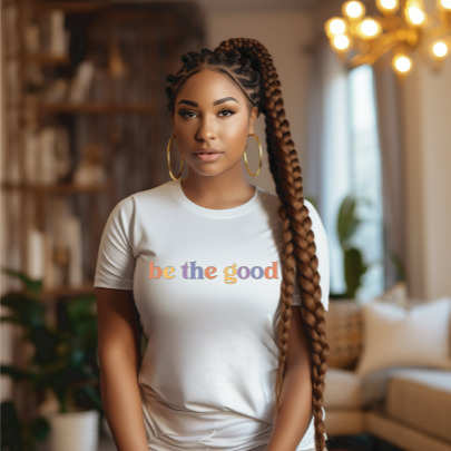 Young woman in short sleevewhite tee with graphic which reads “Be The Good” in colorful letters.