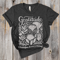 Dark gray t-shirt with white floral design and 'Gratitude Changes Everything' text on a wooden surface