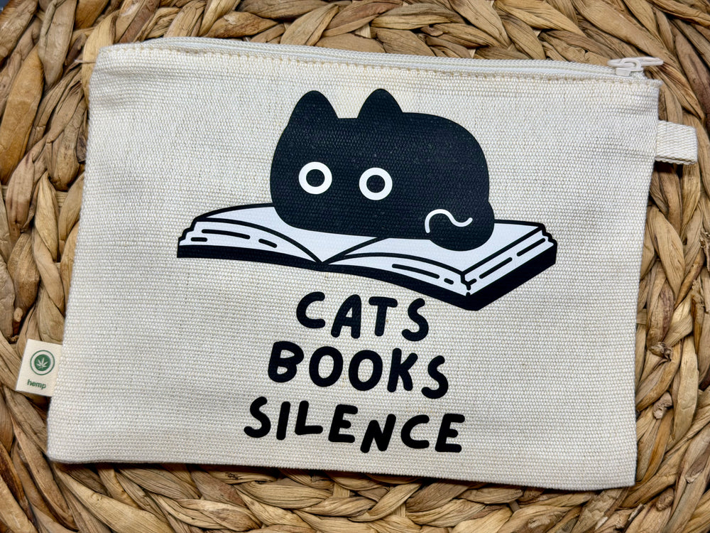 A square canvas pouch with a zipper closure, featuring a black graphic of a cat resting on an open book above the text 'CATS BOOKS SILENCE'. The pouch is laid flat on a woven straw mat.
