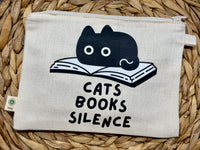 A square canvas pouch with a zipper closure, featuring a black graphic of a cat resting on an open book above the text 'CATS BOOKS SILENCE'. The pouch is laid flat on a woven straw mat.