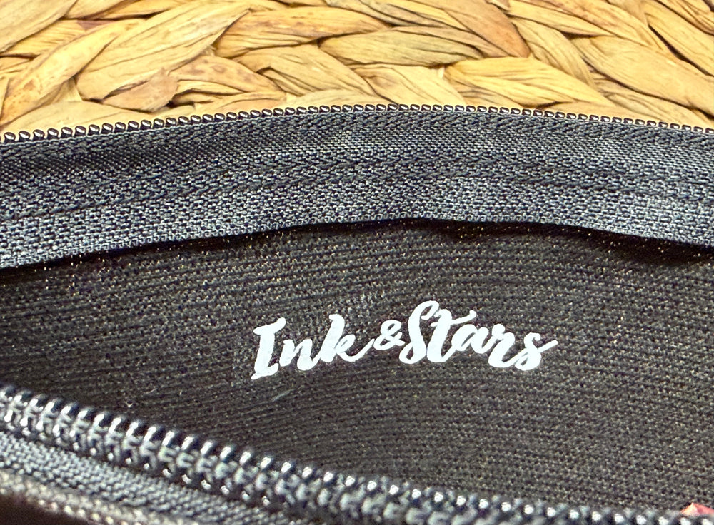 Close-up of a black fabric pouch or case with a white 'Ink&Stars' logo printed on the inside, showing a woven straw surface in the background.