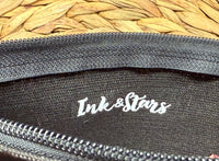 Close-up of a black fabric pouch or case with a white 'Ink&Stars' logo printed on the inside, showing a woven straw surface in the background.