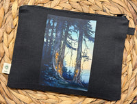 A black canvas zipper pouch featuring a printed vertical art panel of a forest scene with tall trees and warm glowing trunks, displayed on a woven straw mat.