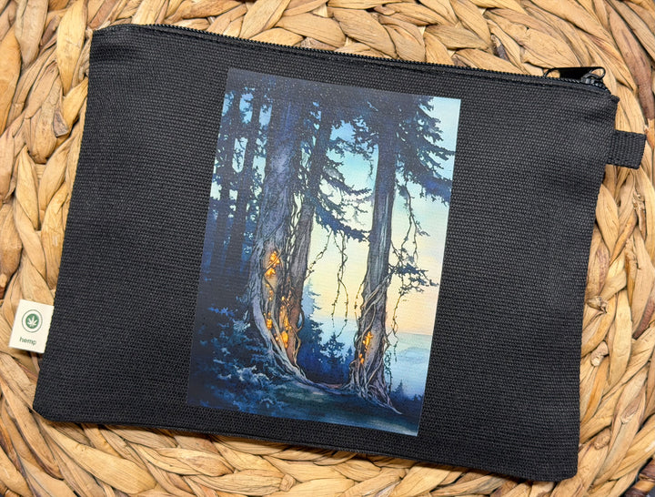 A black canvas zipper pouch featuring a printed vertical art panel of a forest scene with tall trees and warm glowing trunks, displayed on a woven straw mat.