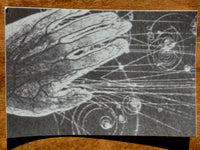 A black and white art print featuring a hand in contact with spiral cosmic patterns, displayed on a wooden surface.