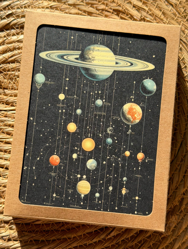 A box of greeting cards featuring a retro cosmic system illustration with planets and stars on a dark background, placed on a woven straw surface.
