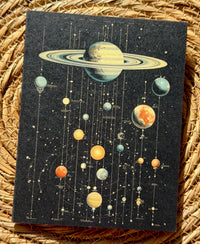 A greeting card featuring a retro-style celestial system with planets, stars, and orbital lines on a dark background, displayed on a woven straw mat.