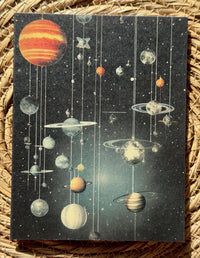 A rectangular art print or card featuring a retro-style solar system illustration with hanging planets and stars on a dark starry background, placed on a woven straw mat.