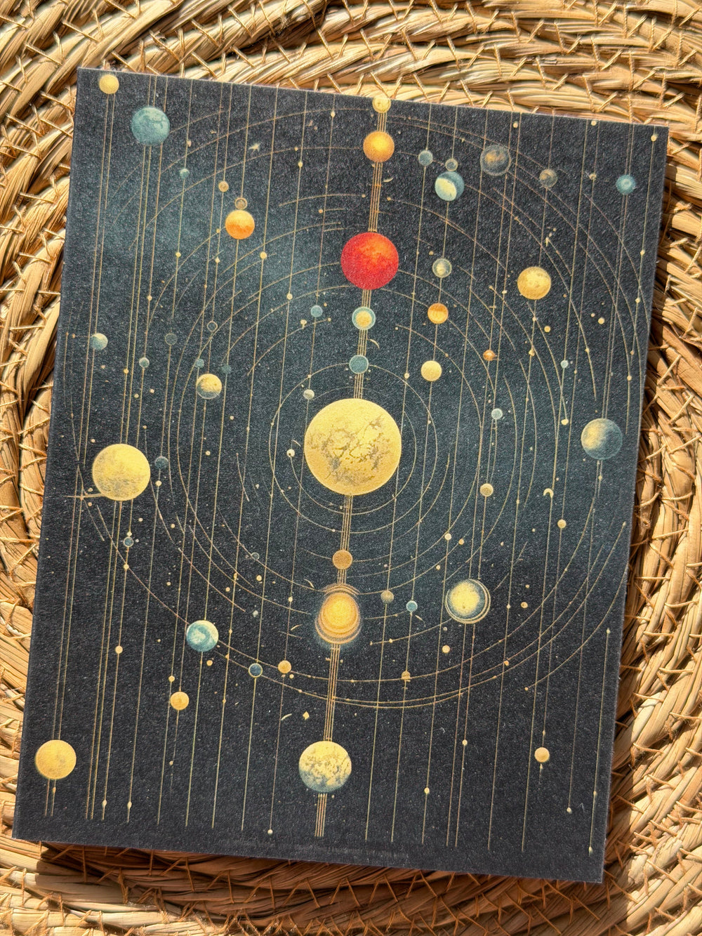 A rectangular art print featuring a stylized solar system with planets and orbits on a dark navy background, placed on a woven straw mat.
