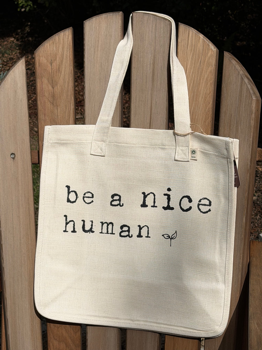 An off-white canvas tote bag with black printed text reading 'be a nice human' and a small leaf graphic, resting on a wooden Adirondack chair outdoors.