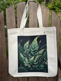 An off-white canvas tote bag with a large rectangular print of green ferns on a black background, displayed on a wooden slatted garden chair.