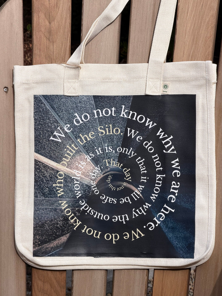 A natural canvas tote bag with a large printed graphic on the front, featuring a circular spiral of white text over a dark, moody background. The bag is photographed hanging on a wooden slatted bench.