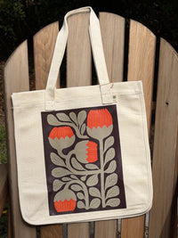 A natural hemp-canvas tote bag with a large botanical print of orange and gray flowers on a dark background, displayed on a wooden Adirondack chair.