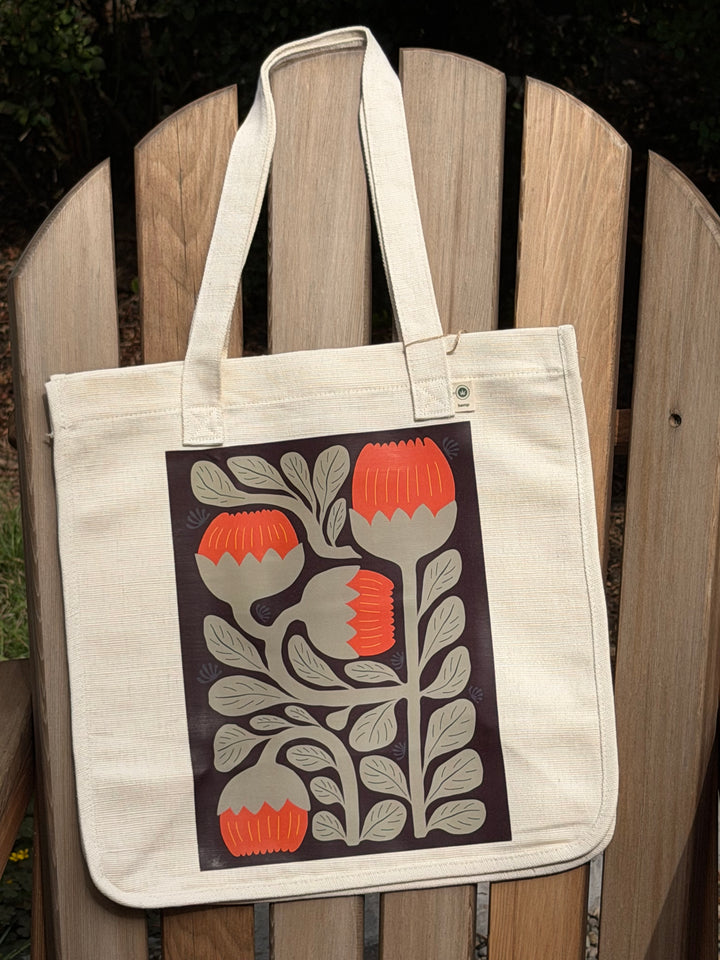 A natural hemp-canvas tote bag with a large botanical print of orange and gray flowers on a dark background, displayed on a wooden Adirondack chair.