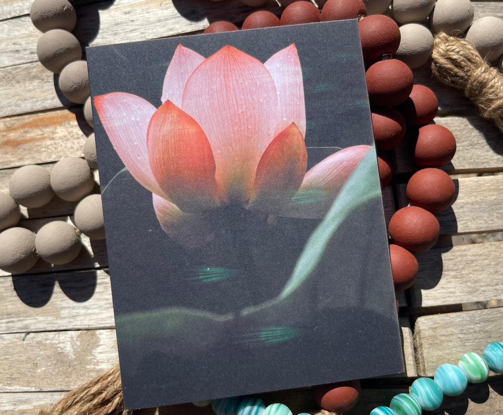 Card with a pink lotus flower on a wooden surface with pebbles and rope.