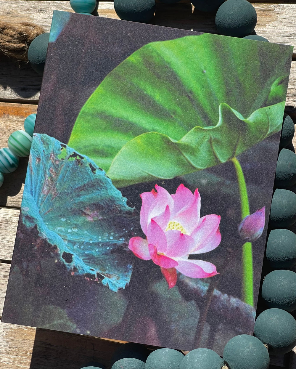 Card with a pink lotus flower and green leaf on a dark background, surrounded by green stones.