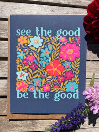 Card with floral design and text 'see the good be the good' on a wooden surface with flowers.