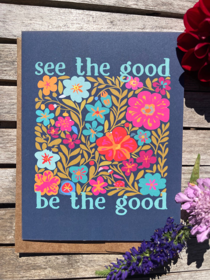 Card with floral design and text 'see the good be the good' on a wooden surface with flowers.