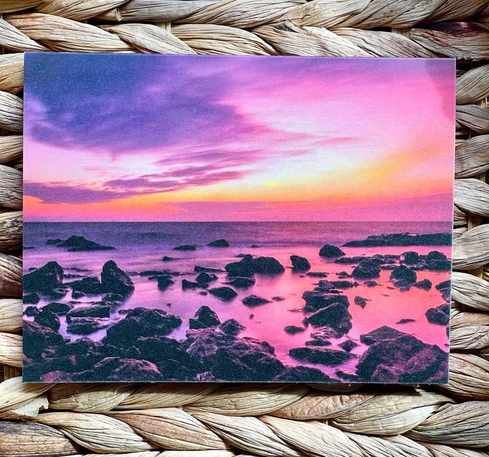 Card with a colorful sunset over a rocky beach, placed on a woven surface.