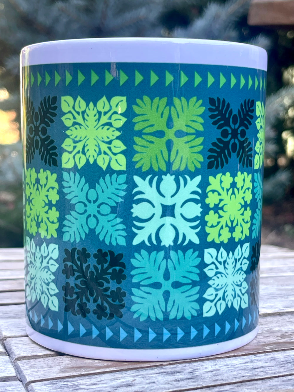 Mug with a geometric Hawaiian quilt pattern of green and blue on a wooden surface