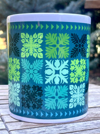 Mug with a geometric Hawaiian quilt pattern of green and blue on a wooden surface