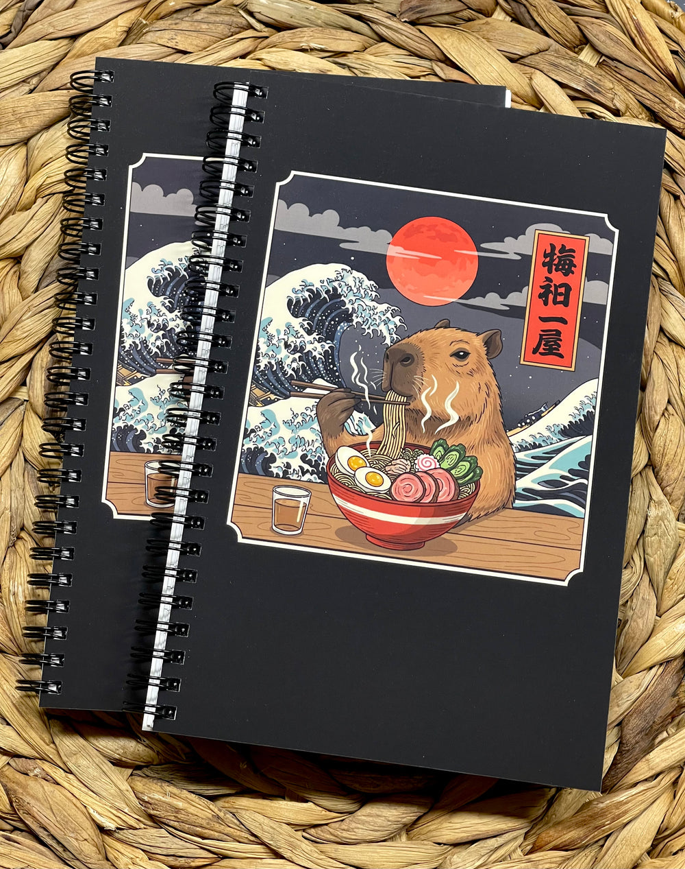 Black notebook with a capybara eating ramen illustration, on a woven surface