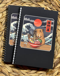 Black notebook with a capybara eating ramen illustration, on a woven surface