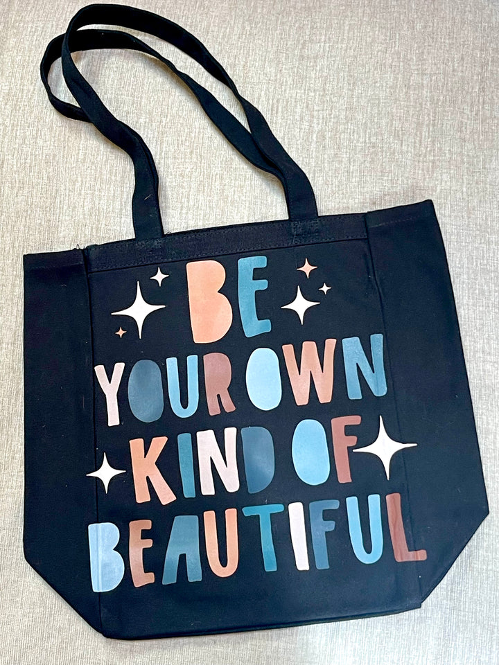 Black tote bag with “Be Your Own Kind Of Beautiful” colorful grphic on front.