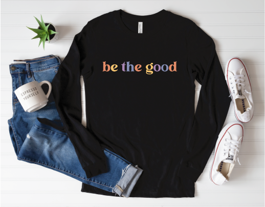 Flat lay of long sleeve black tee with casual life props around it; graphic reads “Be The Good” in colorful letters.