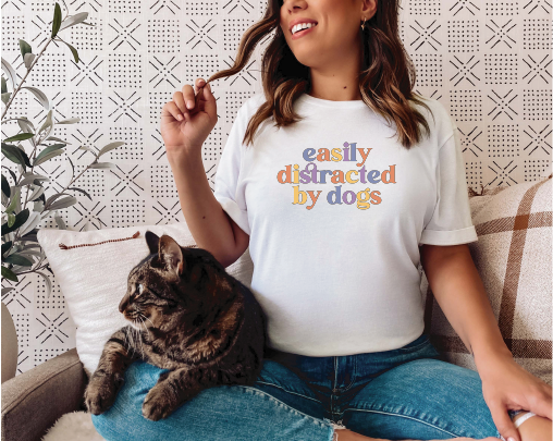 Woman wearing “Easily Distracted By Dogs” white short sleeve tee shirt, petting her cat.