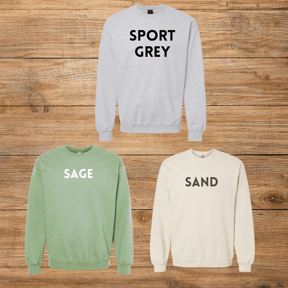 Three crewneck sweatshirts displayed on a rustic wooden background: a light grey sweatshirt with 'SPORT GREY' in black text, a sage green sweatshirt with 'SAGE' in white text, and a sand beige sweatshirt with 'SAND' in black text.