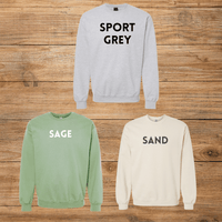 Three crewneck sweatshirts displayed on a rustic wooden background: a light grey sweatshirt with 'SPORT GREY' in black text, a sage green sweatshirt with 'SAGE' in white text, and a sand beige sweatshirt with 'SAND' in black text.