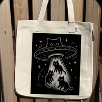 An off-white canvas tote bag with a black graphic print of a UFO catching cats, displayed against a wooden slatted background.