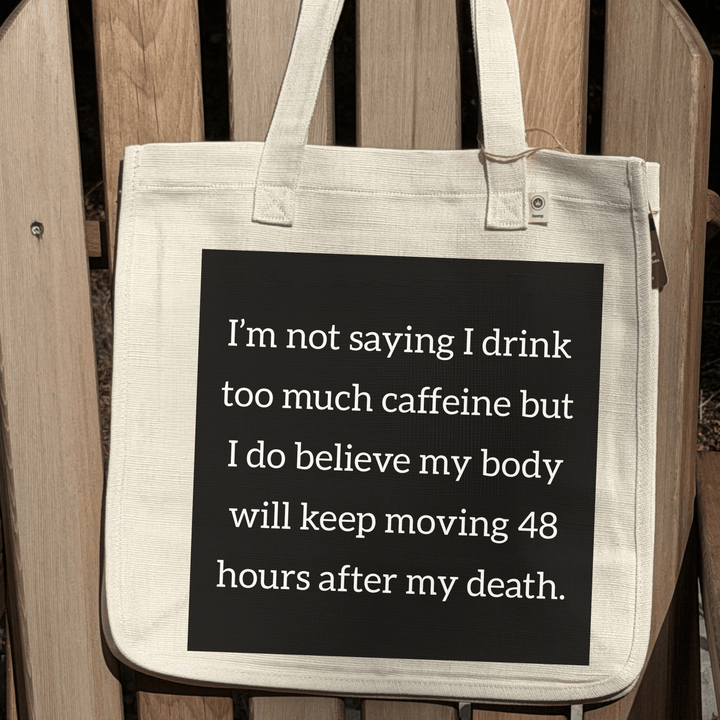 A natural canvas tote bag with a large black square graphic printed with white text about drinking too much coffee, displayed on a wooden slatted bench.